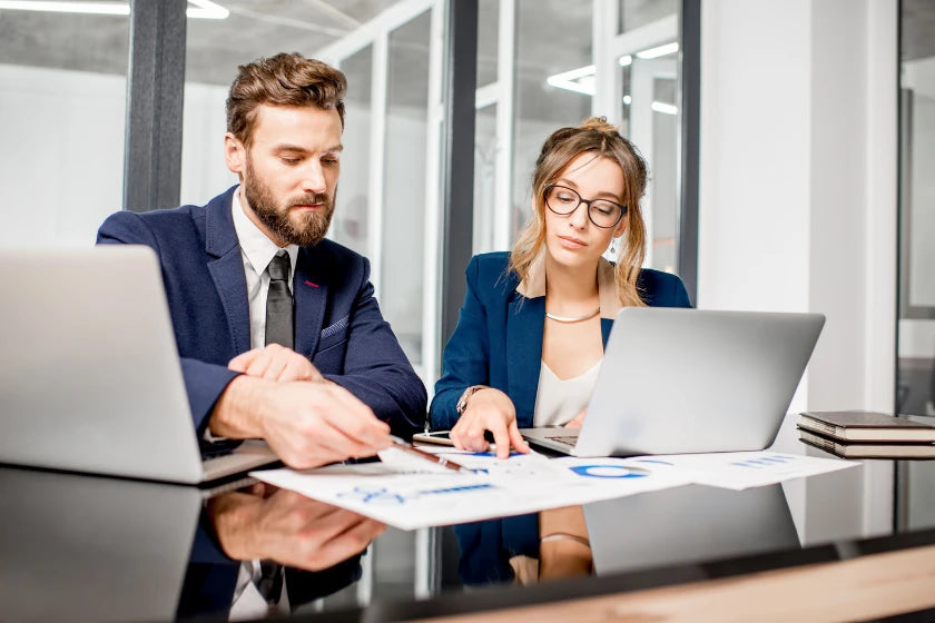 Two business professionals seated at a table, working on laptops and engaged in discussion.
