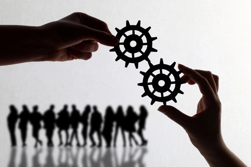 Silhouette of business people with gears, alongside a hand holding a paper cutout of a cogwheel, symbolizing teamwork and innovation.